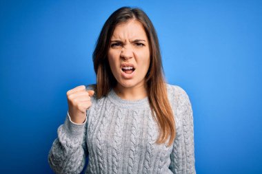 Mavi izole edilmiş arka planda dikilen yünlü süveter giyen genç ve güzel bir kadın öfkeyle bağırırken kızgın ve kızgın bir şekilde yumruğunu kaldırıyor. Öfke ve agresif kavram.