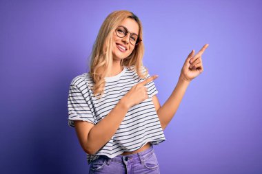 Genç, sarışın, çizgili t-shirt ve gözlük takan, mor arka planda gülümseyen ve kameraya bakan iki eliyle ve parmaklarıyla yüzünü gösteren bir kadın..
