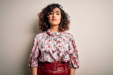 Genç ve güzel kıvırcık Arap kadın çiçekli tişört giyiyor. İzole edilmiş beyaz arka planda duruyor. Yüzünde ciddi bir ifadeyle rahatlamış. Basit ve doğal bir şekilde kameraya bakıyorsun..
