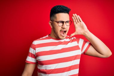 Sıradan çizgili tişört ve gözlük takan genç yakışıklı adam izole edilmiş kırmızı arka planda el ele tutuşarak bağırıyor ve çığlık çığlığa bağırıyordu. İletişim konsepti.