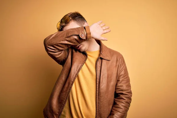 Young handsome redhead man wearing casual leather jacket over isolated yellow background covering eyes with arm, looking serious and sad. Sightless, hiding and rejection concept