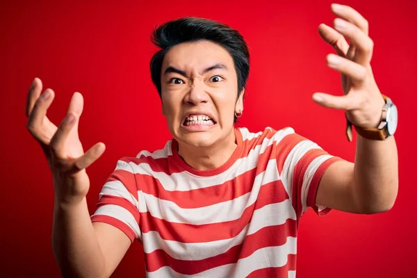 Young handsome chinese man wearing casual striped t-shirt standing over red background Shouting frustrated with rage, hands trying to strangle, yelling mad