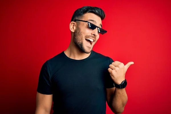 Young handsome man wearing funny thug life sunglasses over isolated red background smiling with happy face looking and pointing to the side with thumb up.