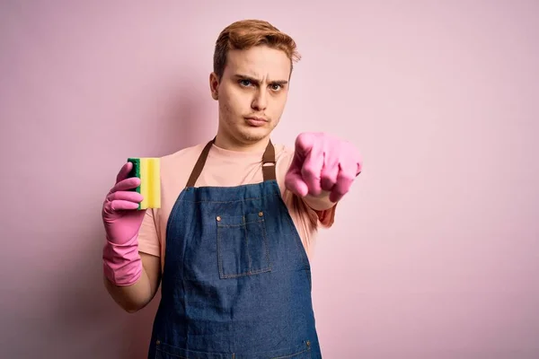 Handsome redhead man doing housework wearing apron and gloves using cleaner scourer pointing with finger to the camera and to you, confident gesture looking serious