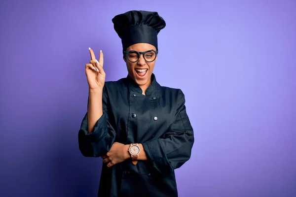 年轻的非洲女厨师身穿炊具制服 头戴紫色背景的帽子 喜形于色地微笑着 用手指在镜头前眨眼示意胜利 第二点 — 图库照片
