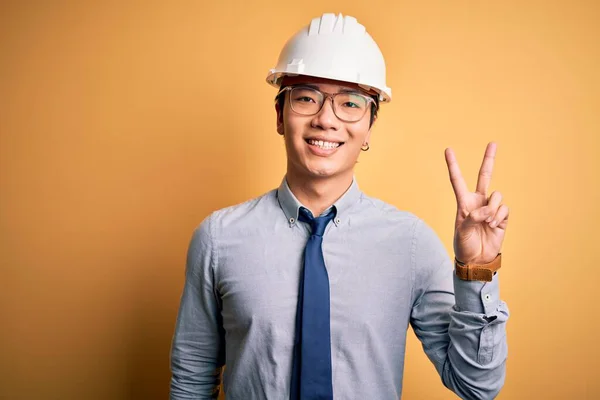 年轻英俊的中国建筑师 戴着安全帽 系在黄色的背景上 笑容满面 在镜头前眨眼示意胜利 第二点 — 图库照片