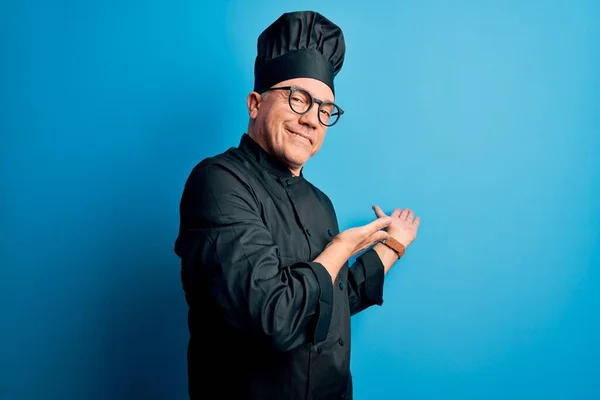 Middle age handsome grey-haired chef man wearing cooker uniform and hat Inviting to enter smiling natural with open hand