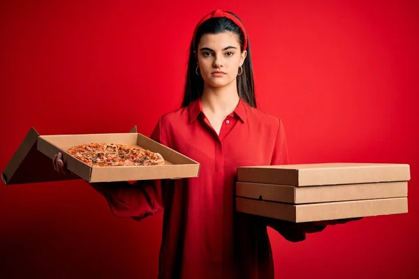 年轻美丽的黑发女子拿着装有意大利披萨的送货盒 背景是红色的 脸上带着自信的表情 脸上带着严肃的表情 — 图库照片