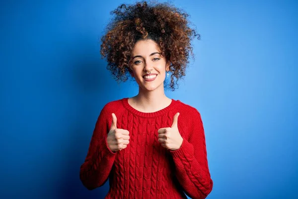 年轻美丽的女人 卷曲的头发 头戴宽松的红色毛衣 穿孔成功的标志 用手做积极的手势 竖起大拇指 微笑着 快乐地 快乐的表情和胜利的姿态 — 图库照片