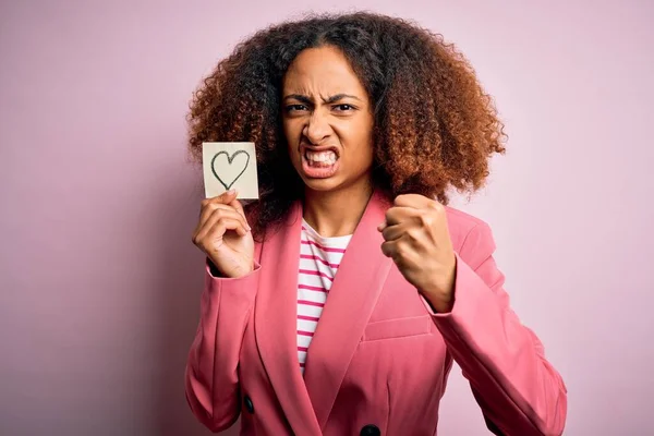 Young african american woman with afro hair holding paper with heart over pink background annoyed and frustrated shouting with anger, crazy and yelling with raised hand, anger concept