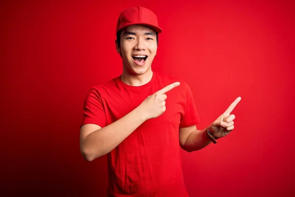 Young handsome chinese delivery man wearing cap standing over isolated red background smiling and looking at the camera pointing with two hands and fingers to the side.