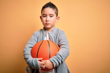 Basketbol topuyla oynayan küçük bir çocuk izole edilmiş sarı arka planda akıllı bir yüz ifadesiyle ciddi ciddi düşünüyor.