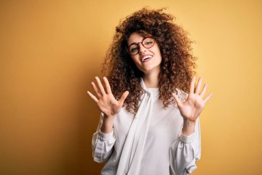 Kıvırcık saçlı, piercing takan, gömlek takan, gözlük takan, on numaralı parmaklarıyla onu gösteren, kendinden emin ve mutlu bir şekilde gülümseyen güzel esmer kadın..