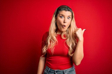 İzole edilmiş kırmızı arka plan üzerinde duran gündelik tişört giyen genç ve güzel sarışın kadın şaşırmış bir şekilde elini kenarı işaret ediyor, ağzı açık şaşkın bir şekilde ifade ediyor..