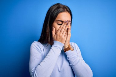 Genç, güzel, esmer bir kadın mavi arka planda dikilip ağlarken suratını ellerle kapatan sıradan bir kazak giyiyor. Depresyon kavramı.