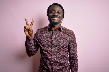 Genç, yakışıklı, Afro-Amerikalı bir adam pembe arka planda dikilmiş gülümsüyor ve parmaklarını zafer işareti yaparken gösteren kameraya bakıyor. İki numara..
