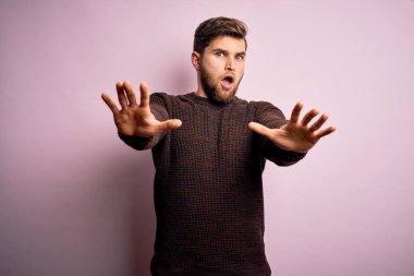 Sakallı ve mavi gözlü genç bir adam pembe arka planda sıradan bir süveter giyiyor. Avuç içi hareketlerini durduruyor. Öfkeli ve hüsran dolu bir ifade takınıyor.