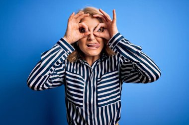 Orta yaşlı, sarışın, sıradan çizgili tişört giyen, mavi arka planda duran güzel bir kadın. Dürbün gibi güzel hareketler yapıyor. Gözleri parmaklarından bakıyor. Çılgın ifade.