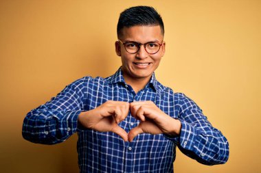 Sarı arka planda gündelik tişört ve gözlük takan genç, yakışıklı Latin bir adam. Aşkla gülümsüyor, elleriyle kalp sembolü yapıyor. Romantik konsept.