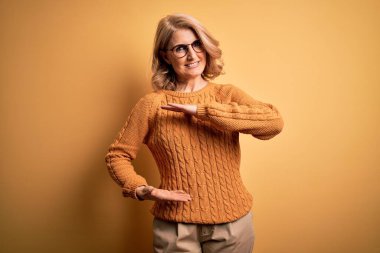 Orta yaşlı, güzel, sarışın bir kadın sarı arka planda gözlük ve süveter takıyor. Elleri büyük ve büyük boy işareti, ölçü sembolü gösteriyor. Kameraya bakıp gülümsüyor. Ölçüm konsepti.