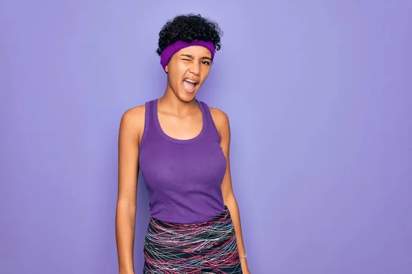 美しいアフリカ系アメリカ人アフロ女性身に着けていますカジュアルスポーツウェア以上紫の背景ウィンク見ますカメラでセクシー式 陽気で幸せな顔 — ストック写真