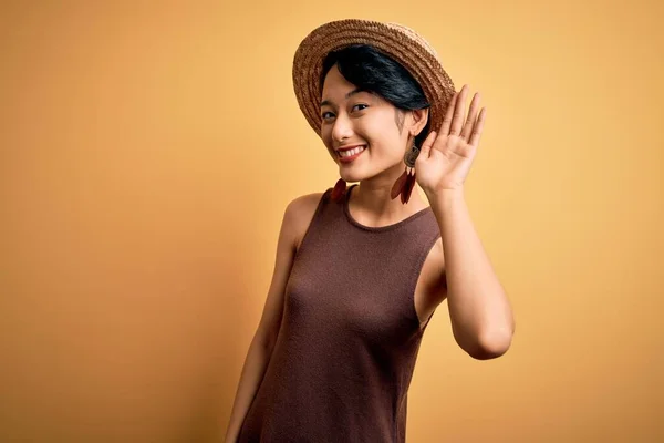 年轻美丽的亚洲女孩穿着休闲装 头戴一顶帽子 头戴黄褐色背景的帽子 面带微笑 两手相对地听着流言或流言蜚语 失聪的概念 — 图库照片