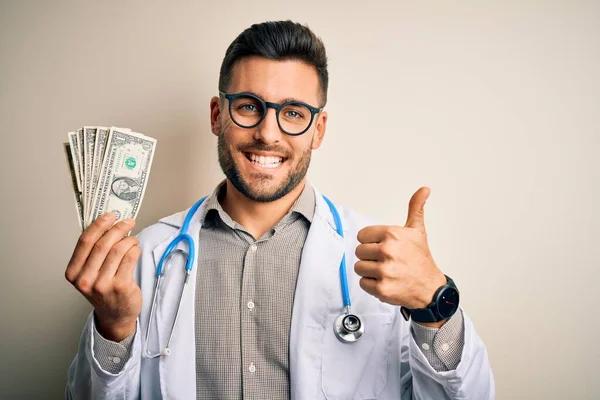 大きな笑顔で幸せな孤立した背景の上に1ドル紙幣の束を保持若い医師の男はOkサインをし 親指を指で開くと 優れたサイン — ストック写真