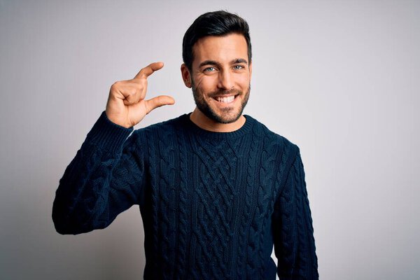 Young handsome man with beard wearing casual sweater standing over white background smiling and confident gesturing with hand doing small size sign with fingers looking and the camera. Measure concept.