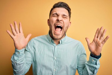 Mavi gözlü genç iş adamı sarı arka planda zarif yeşil gömlek giyerek çılgın ve başarıyı kollarını kaldırarak ve gözleri kapalı heyecanla haykırarak kutluyor. Kazanan konsept