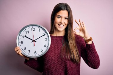 Geri sayım yapan genç güzel bir kız. Pembe arka planda büyük bir saat tutuyor. Tamam işareti yapıyor, mükemmel bir sembol.