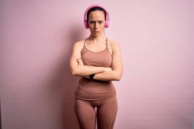 Spor kıyafetleri giyen, kulaklık takan, kuşkucu ve gergin, yüzünde çapraz kollu tasvip etmeyen bir ifade dinleyen genç bir fitness kadını. Negatif kişi.