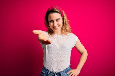 İzole edilmiş pembe arka plan üzerinde sıradan tişörtler giyen genç ve güzel sarışın bir kadın selamlama ve hoş geldin demek için dostça el sıkışmalar yapıyor. Başarılı bir iş..