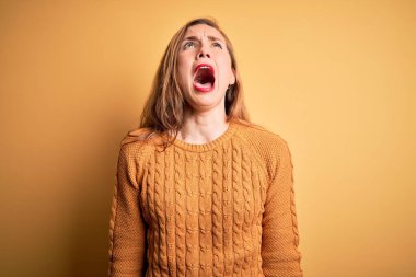 Sarı arka planda sıradan bir süveter giyen genç ve güzel bir kadın sinirli ve öfkeli bir şekilde bağırıyor. Öfkeyle bağırıyor. Öfke ve agresif kavram.