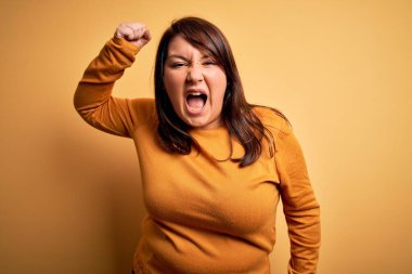 Güzel esmer ve cüsseli bir kadın izole edilmiş sarı arka plan üzerine gündelik süveter giyer kızgın ve kızgın bir şekilde yumruğunu kaldırır ve öfkeyle bağırır. Öfke ve agresif kavram.
