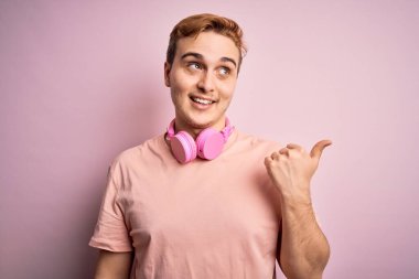 Genç, yakışıklı, kızıl saçlı, müzik dinleyen, pembe arka planda kulaklık takan, gülümseyen, gülümseyen ve baş parmağıyla yana bakan bir adam..