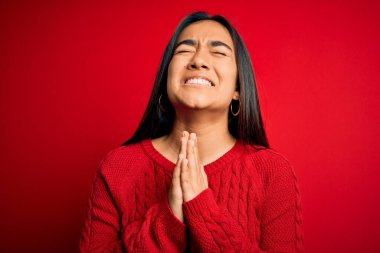 Genç ve güzel Asyalı kadın, günlük süveter giyip izole edilmiş kırmızı arka planda duruyor. Elleriyle yalvarıyor ve dua ediyor. Yüzünde umut ifadesi var. Yalvarıyorum..