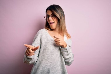 Genç, güzel esmer kadın pembe arka planda rahat süveter ve gözlük takıp mutlu ve komik yüzlü parmakları kameraya doğrultuyor. İyi enerji ve titreşimler.