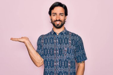 Genç, yakışıklı, bohem, pembe arka planda hippi tarzı giyinmiş neşeli bir sunum yapan ve avuç içi kameraya bakan biri..