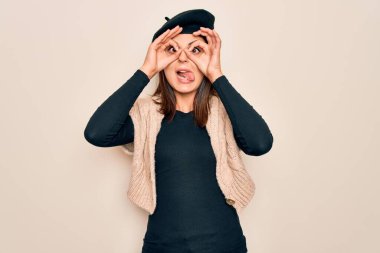 Genç, güzel esmer kadın, beyaz arka plan üzerine Fransız beresi takmış dürbün gibi jest yapıyor, gözleri parmaklarından bakıyor. Çılgın ifade.