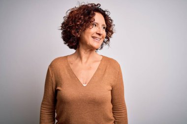 Orta yaşlı güzel kıvırcık saçlı kadın izole edilmiş beyaz arka plan üzerine günlük süveter giyip yüzünde gülümseme, doğal bir ifade ile yan yana bakıyor. Kendinden emin gülen.