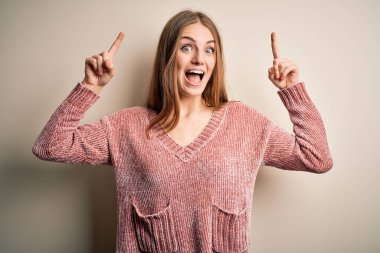 Genç, güzel, kızıl saçlı, pembe bir süveter giyen, izole edilmiş beyaz bir arka plan giyen, şaşkın, şaşkın, parmaklarıyla işaret eden ve kollarını kaldıran bir kadın..