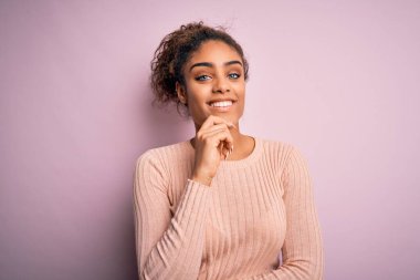 Genç ve güzel Afro-Amerikan kızı pembe arka plan üzerinde sıradan bir süveter giyerek kameraya bakarken kollarını kavuşturup, ellerini çenesine kaldırarak gülümsüyor. Olumlu düşünüyorum..