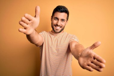 Sarı arka planda sıradan çizgili tişört giyen sakallı yakışıklı genç adam kameraya sarılmak için gülümseyerek bakıyor. Mutluluğu kucaklayan neşeli bir ifade..