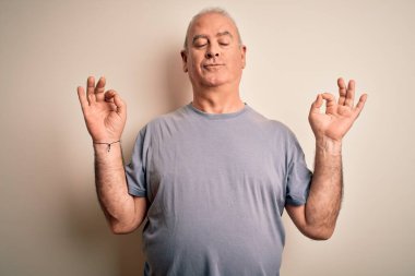 İzole edilmiş beyaz arka plan üzerinde duran beyaz tişörtlü, orta yaşlı yakışıklı adam rahatla ve gözleri kapalı, parmaklarıyla meditasyon hareketi yaparken gülümse. Yoga konsepti.
