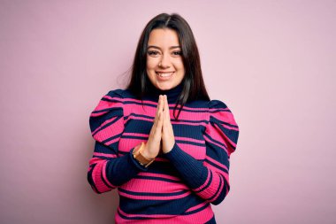 Genç, esmer, zarif bir kadın pembe tişörtlü, soyutlanmış arka planda dua ediyor, elleriyle birlikte af diliyor kendinden emin bir şekilde gülümsüyor..