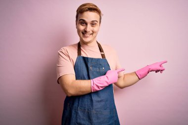 Genç, kızıl saçlı, ev işi yapan, önlüklü, eldivenli, gülümseyen ve kameraya bakan, iki eliyle ve parmaklarıyla yüzünü gösteren bir temizlikçi..