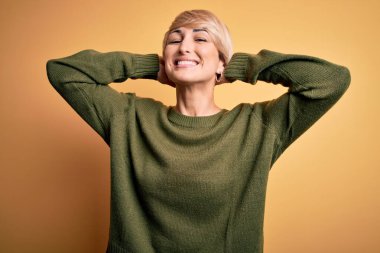 Modern kısa saçlı genç bir kadın sarı arka planda sıradan bir süveter giyiyor rahatlıyor ve esniyor, kolları ve elleri başının arkasında ve boynunun arkasında mutlu bir şekilde gülümsüyor.