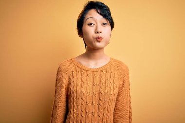 Genç, güzel Asyalı bir kız gündelik süveter ve diyabet giyip sarı arka planda dikilmiş komik suratlı, pofuduk yanakları şişiriyor. Havayla şişmiş ağız, çılgın bir ifade..