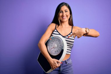 Genç, güzel esmer kadın, izole edilmiş mor arka planda tartı makinesini tutuyor. El ve parmağıyla işaret ederek çok mutlu.