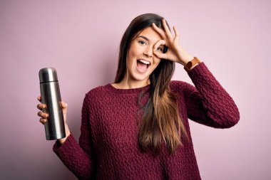 Su dolu termosu olan genç ve güzel bir kız mutlu yüzüyle pembe arka planda duruyor. El ele verip parmaklarından bakıyor.